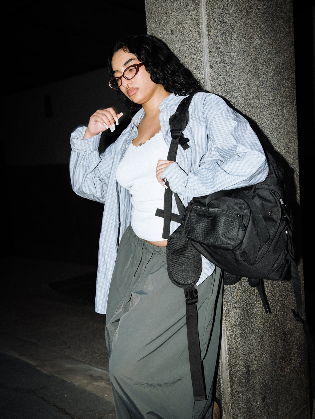 A woman wearing a light blue and white striped button-up shirt, a white tank top, and loose dark green pants, carrying a black crossbody bag with a cross detail on the strap. She is standing outdoors at night next to a textured gray stone pillar.