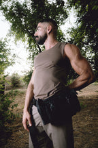 A muscular man with a beard wearing a fitted tan sleeveless shirt and khaki pants, standing outdoors in a wooded area with a black tactical belt pouch at his waist.