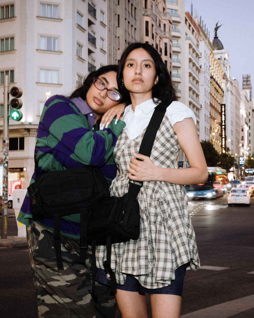 Two young women posing together on a city street at dusk. The woman on the left wears a green and purple striped hoodie, a purple top, and camouflage pants, carrying a black shoulder bag.