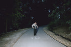 A man in a light-colored shirt and dark trousers stands on a dark, curving road surrounded by trees at night, with the number 10 painted on the road.